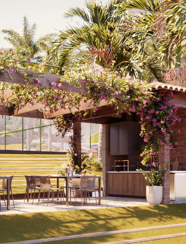 Churrasqueira aberta com gazebo de madeira e flores rosas. Mesas, churrasqueira de tijolinho e balcão de suporte completam o ambiente.